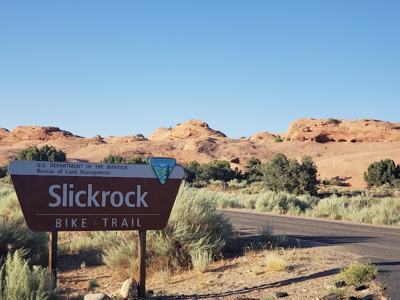 Arches National Park: Slickrock Bike Trail sign beside a dusty road, with red rock formations, desert shrubs, and a clear blue sky.