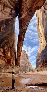 Hiker stands beside a muddy pool in a narrow slot canyon with towering red rock walls and a blue sky above, Arches National Park.