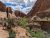 Hikers with backpacks walk a dusty trail through red sandstone canyon walls and green shrubs in Arches National Park.