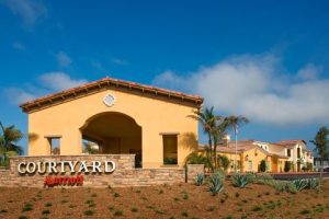 Lodge-style courtyard building with arched entry and warm yellow walls, set in a stone plaza with palm trees.