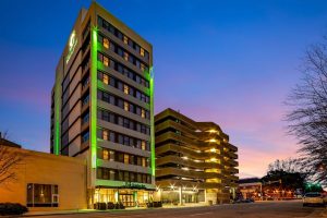 Downtown Columbia hotel exterior at dusk, green neon lights outlining the modern facade and warmly lit lobby.