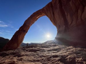Corona Arch frames a sunlit desert panorama in Canyonlands National Park, with rugged sandstone and a blue sky.