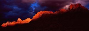 Sunset-lit sandstone cliffs in Zion National Park at dusk, with dark stormy clouds overhead and a fiery orange glow along the ridges.