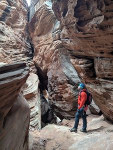 Hiker in a red jacket and helmet stands in a towering slot canyon at The Narrows, Zion National Park.