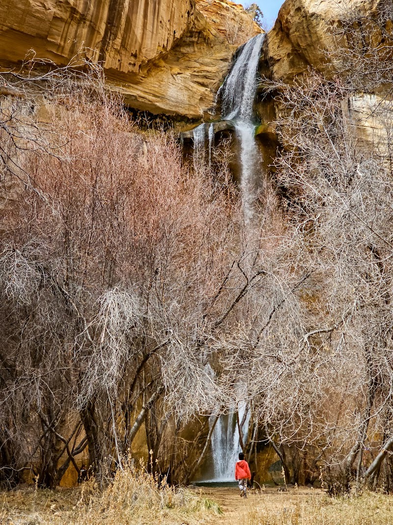 Hiker in a red rain jacket stands before a tall waterfall in a slot canyon national park