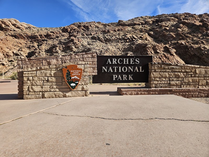 Stone entrance with a black Arches National Park sign and a National Park Service shield set against red cliff rocks.