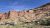 Arches National Park: red sandstone cliffs rise over a small visitor center with parked cars, desert shrubs, under a blue sky.