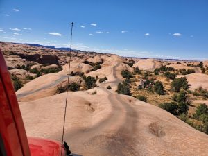 View from a red 4x4 along a winding sandy trail over smooth orange rock fins with scattered trees in Arches National Park.
