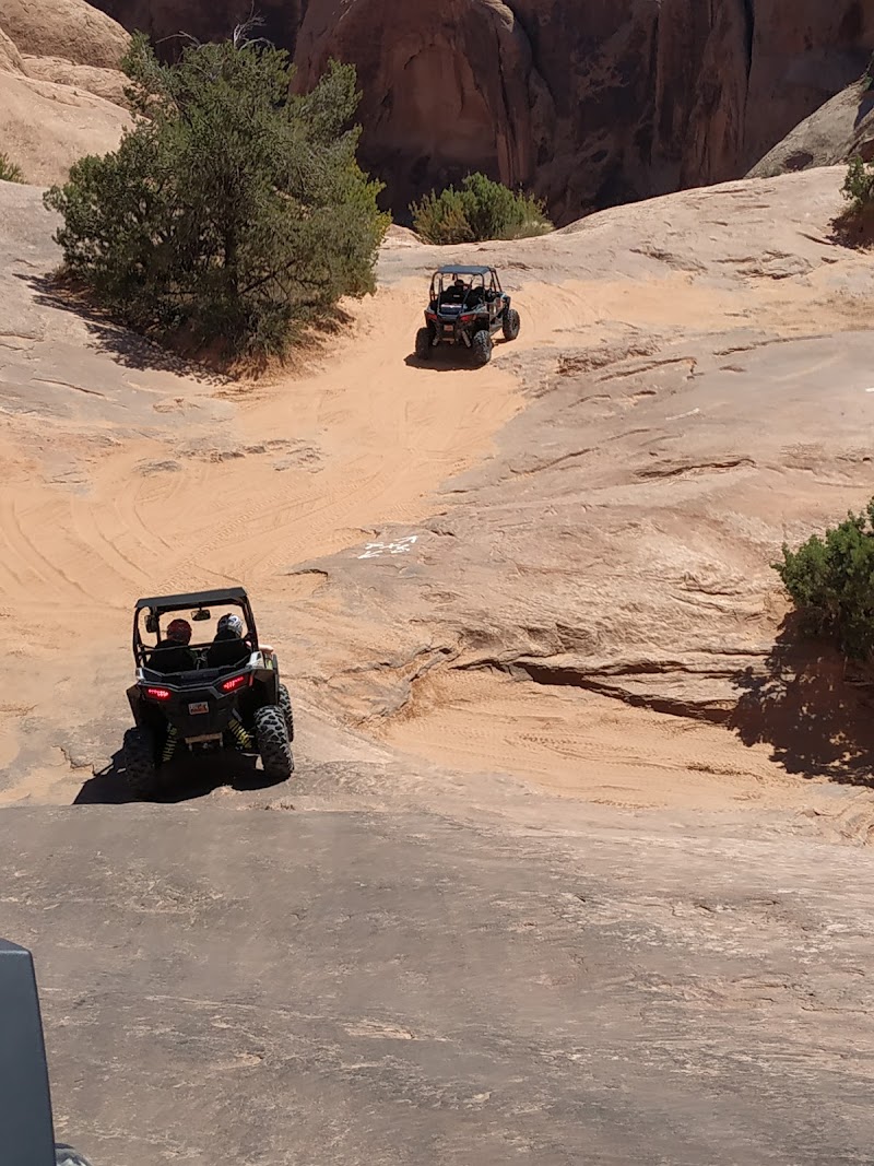 Two side-by-side off-road UTVs navigate a sandy, rocky trail amid red arches in Arches National Park.