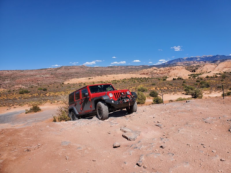 Red Jeep Wrangler on a rocky 4x4 trail in Arches National Park amid desert scrub and distant mesas under a clear blue sky.