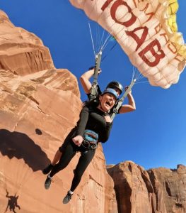 Tandem skydiving over red sandstone cliffs in Arches National Park, two riders in black suits under a bright parachute.