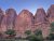Red sandstone cliffs rise above a rocky base with green trees and a clear blue sky, at Arches National Park.