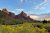 A triangular sandstone mountain overlooks green and yellow foliage. A cloudy blue sky is overhead.