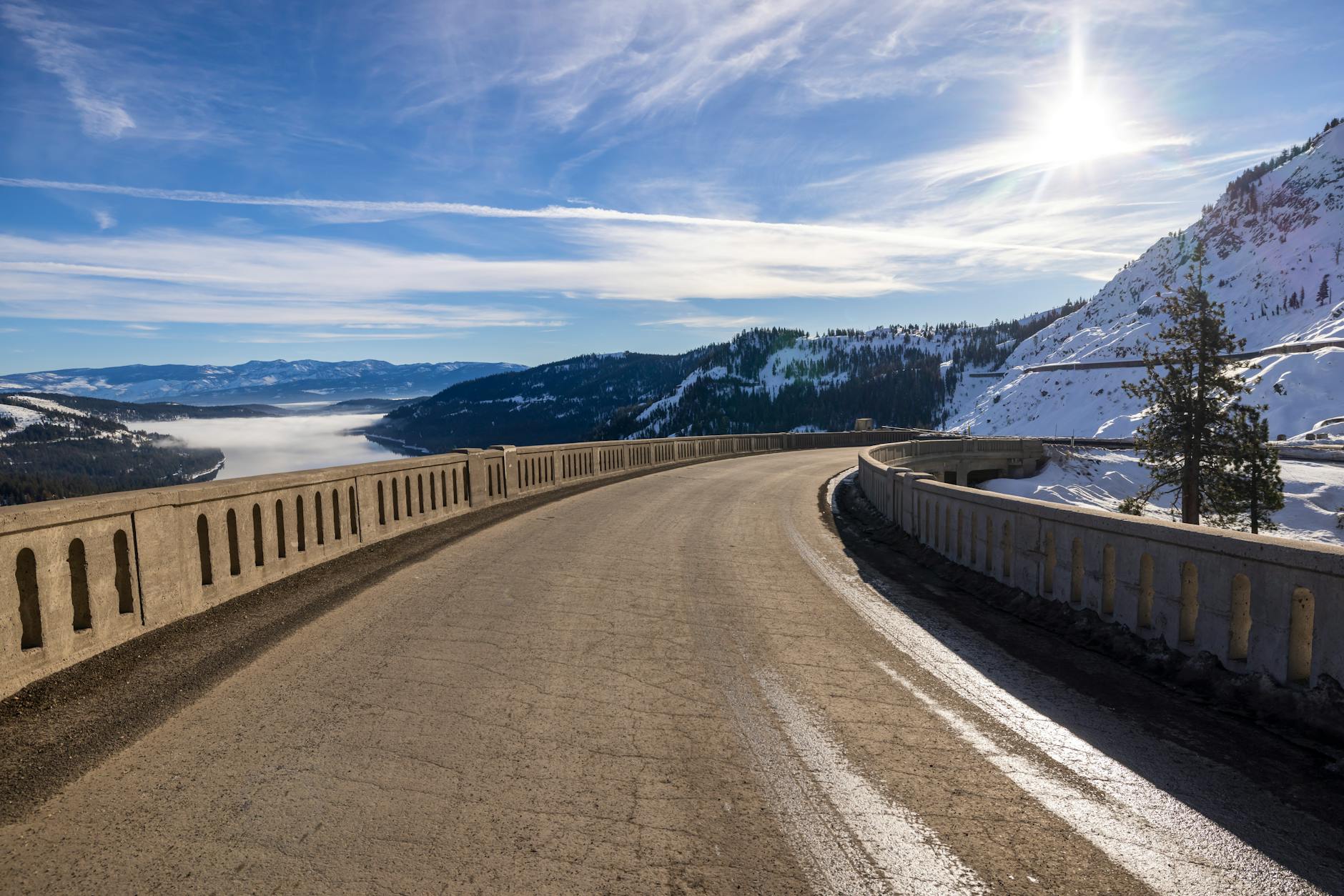 Snowy road curving through Donner Pass with a sunny sky and mountainous backdrop.