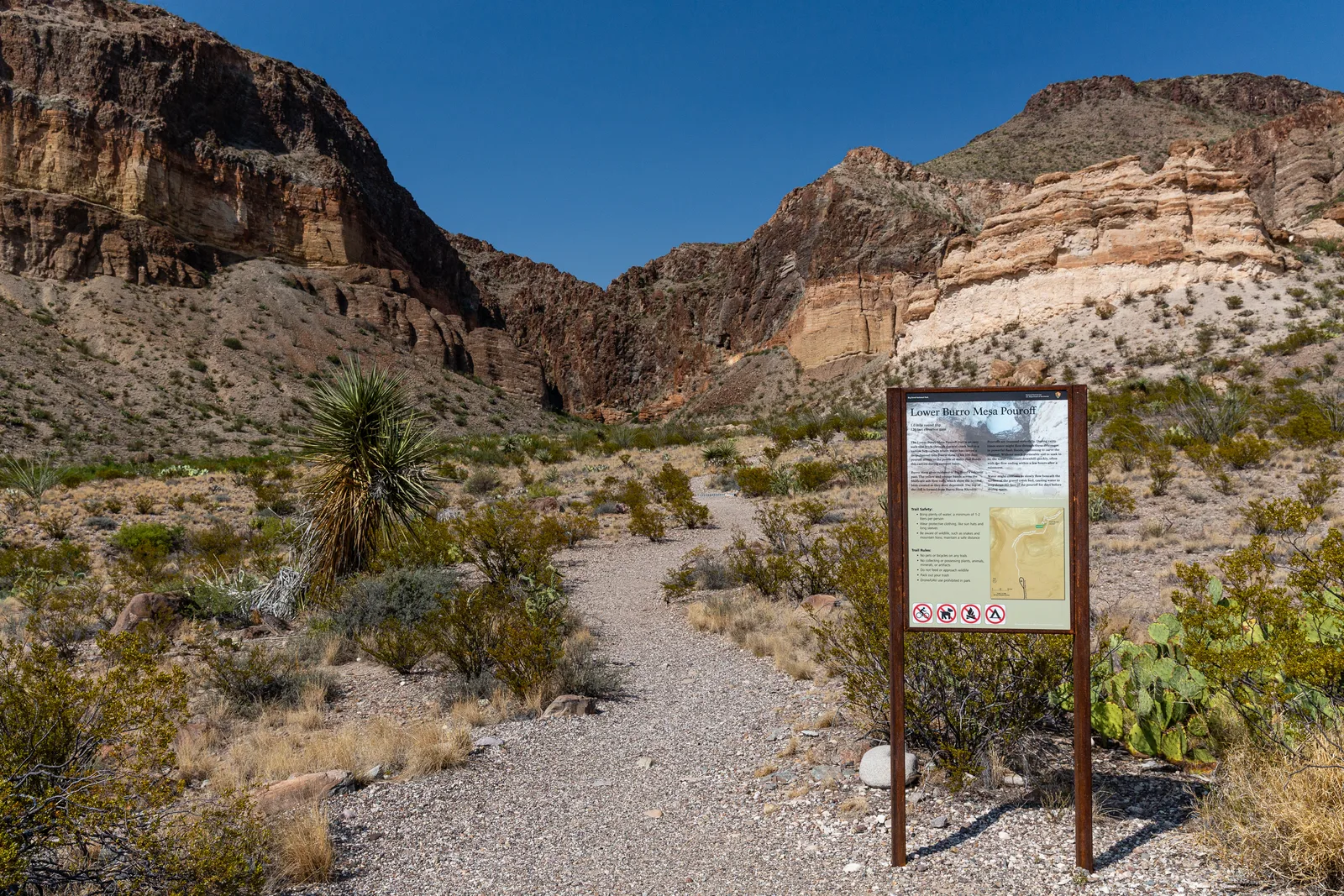 A gravel path starts at a trailhead sign and winds back to colorful igneous cliffs.