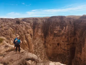 Two hikers peer over the edge of a high limestone cliff.