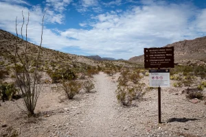 A metal sign with trail information sits beside a dirt path heading into the mountains.