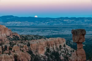 A full moon rises in a blue and purple sky over the red rock canyon.