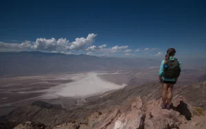 A hiker stands on a rocky ridge looking away from the camera out on a vast white desert valley.