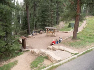 Several people are sitting in a rock-lined open trailhead area, with a footpath on the right.