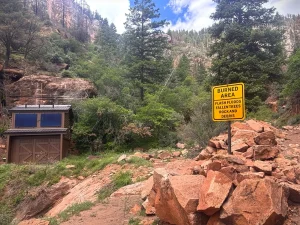 Composting toilet by a trail in forested area with sign warning hikers of potential hazards.