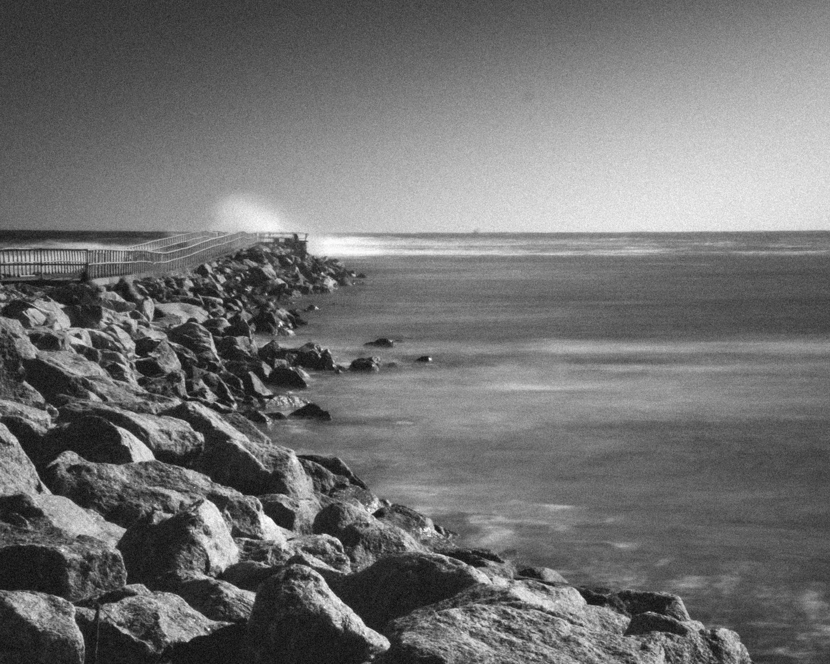 A striking black and white view of a rocky seashore with a pier stretching into the ocean.