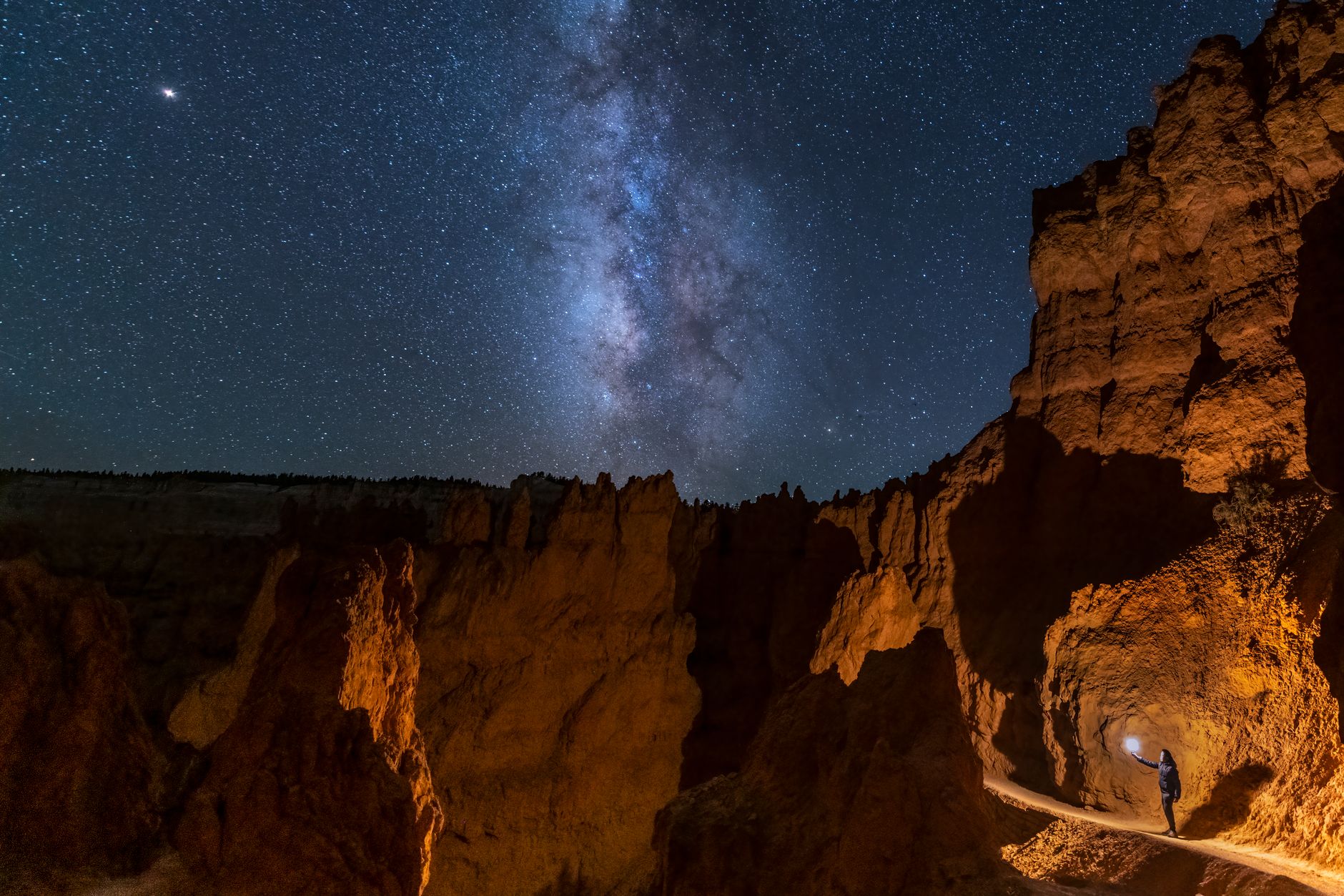 A lone traveler explores Bryce Canyon at night, illuminated by the Milky Way in a clear starry sky.