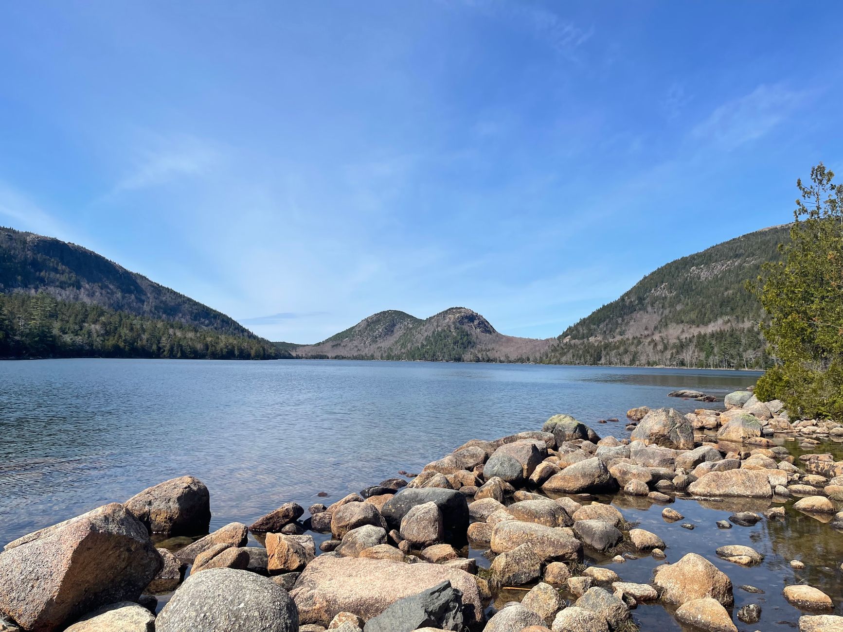 Rocky shoreline at Acadia National Park with clear waters and distant hills under a sunny sky.