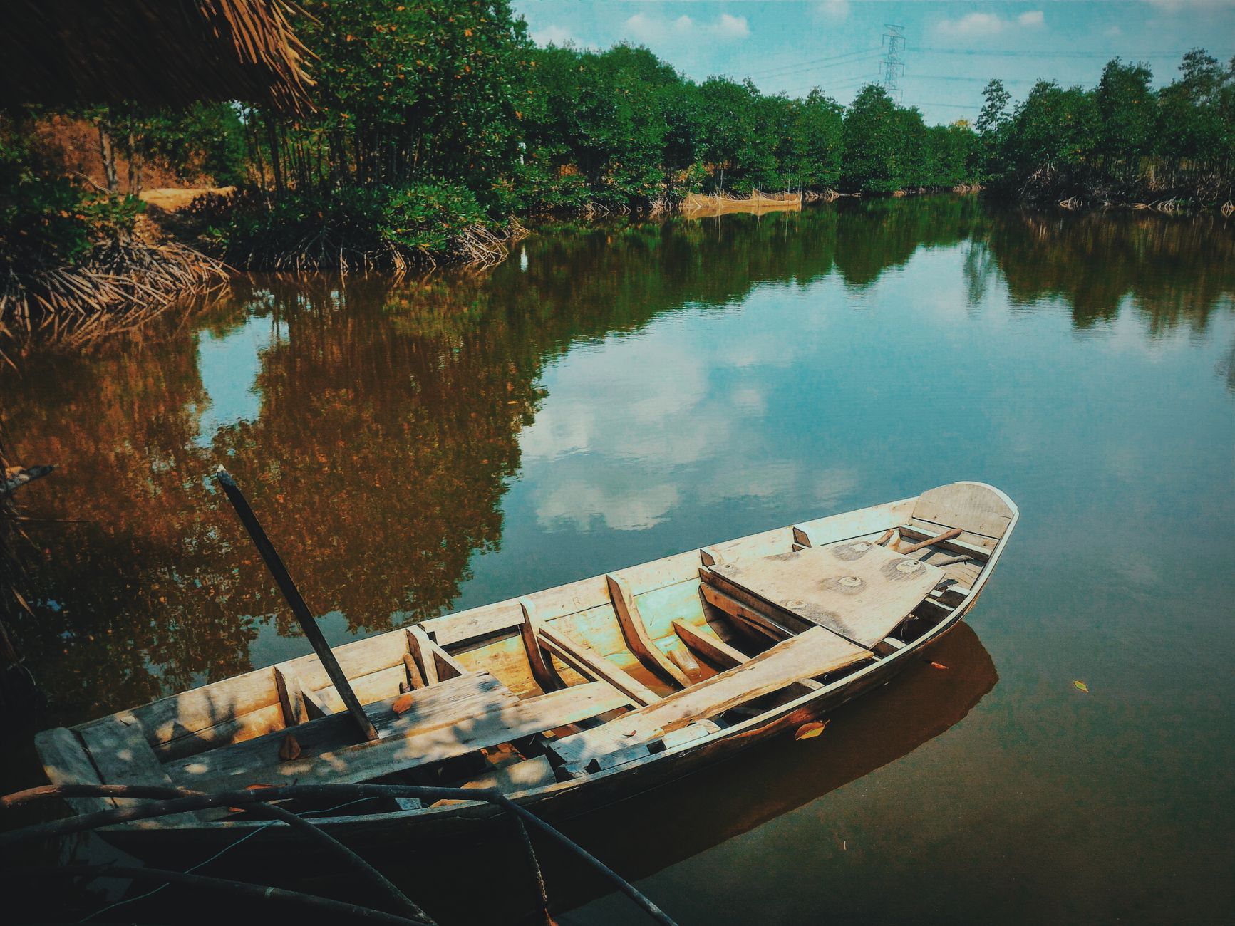 A peaceful scene of a wooden boat reflecting on a calm river surrounded by lush greenery under a clear blue sky.