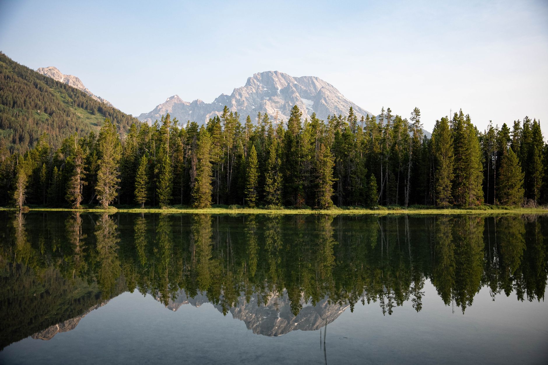 Beautiful reflection of the Grand Tetons in a serene lake surrounded by evergreen trees in Jackson, Wyoming.