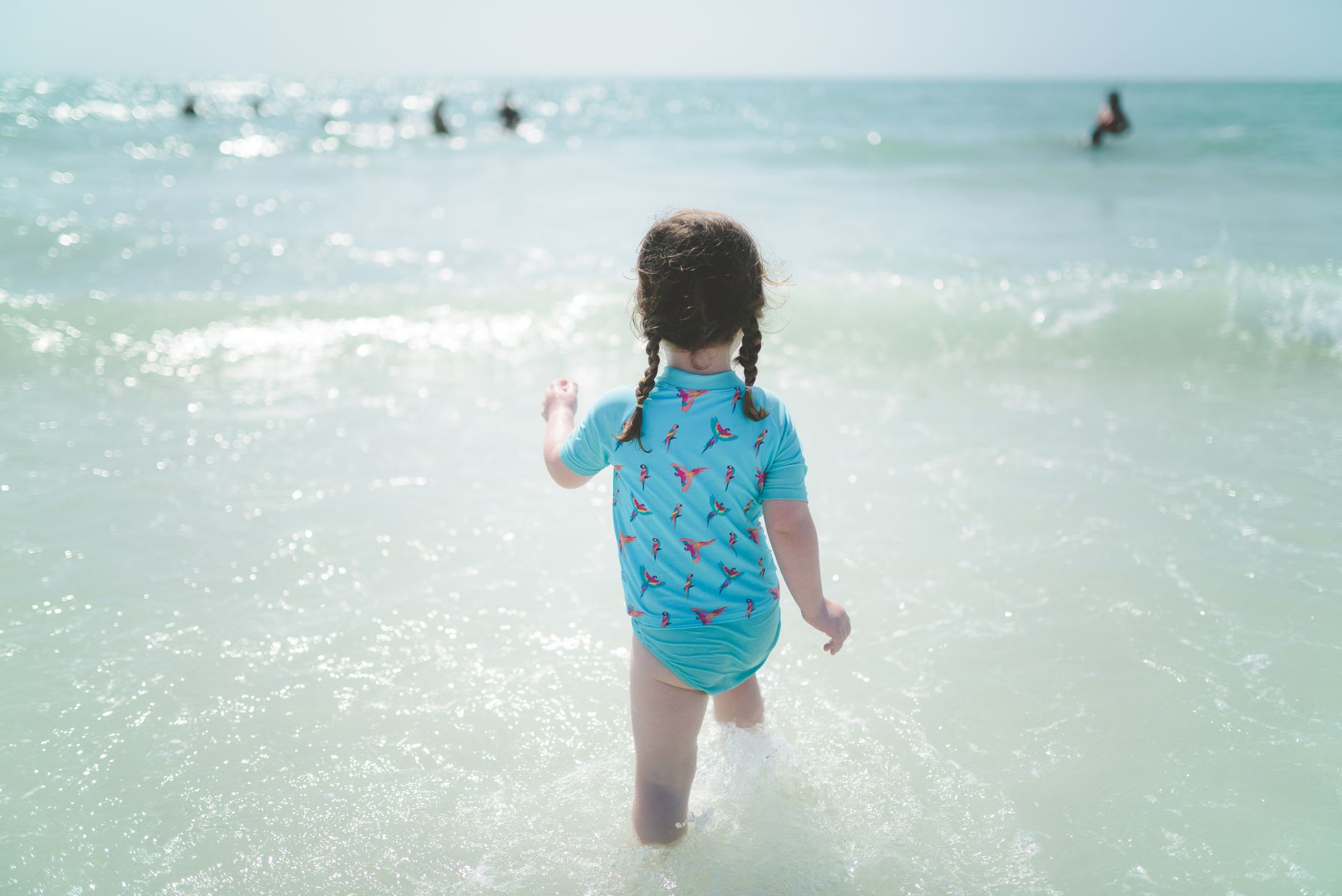 A young girl with braided hair walks into the ocean waves under the sunny sky.