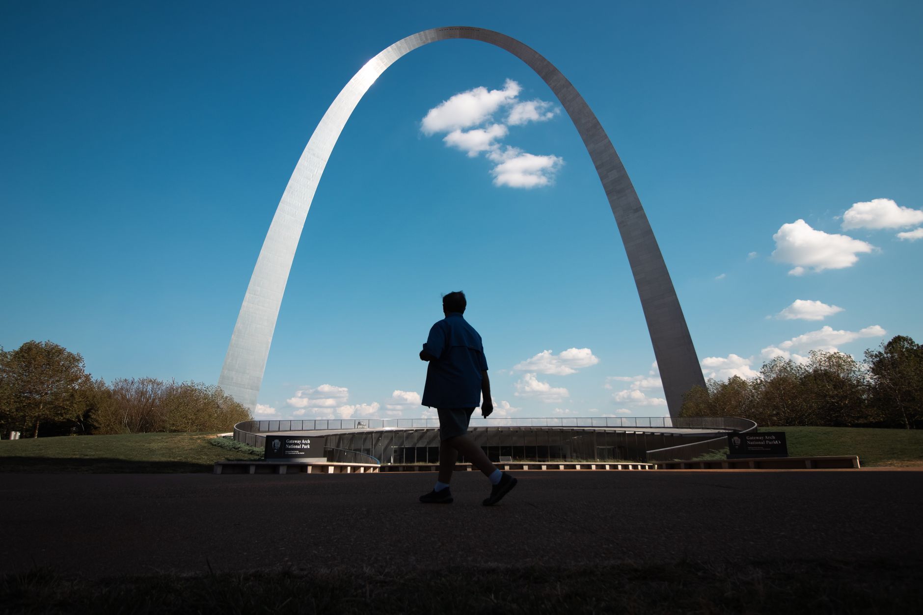 Silhouette of a person walking by the iconic Gateway Arch under a clear blue sky.