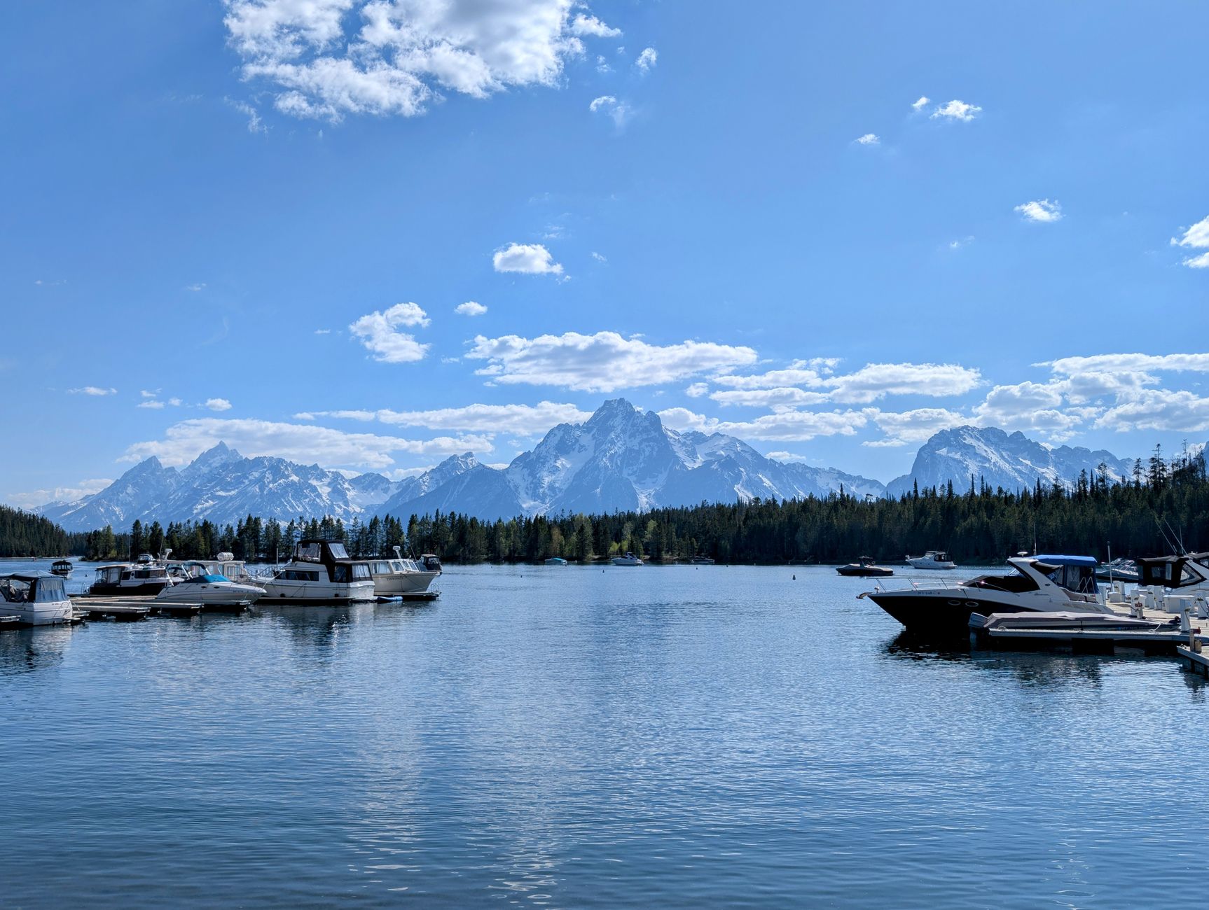 Tranquil marina scene with boats on a lake, framed by the majestic Grand Tetons under a clear blue sky.