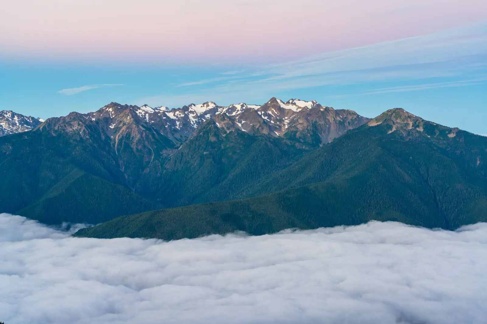 Majestic mountain range with snow-capped peaks above clouds during dawn light, showcasing natural beauty.